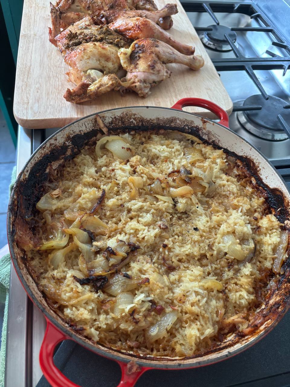 A wide red Le Creuset casserole pan filled with baked rice and caramelised onion wedges with a golden, slightly charred crust around the edges, sitting on a gas stovetop. In the background, a carved spatchcock roast chicken rests on a wooden chopping board, its skin dark and herb-crusted.