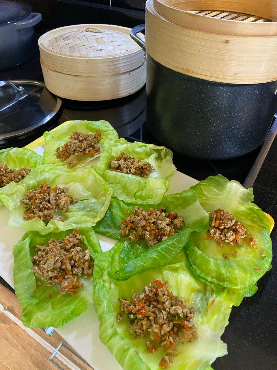 Around ten blanched cabbage leaves arranged on a white chopping board, each filled with a spoonful of cooked mince mixed with grated carrot, red pepper and mushrooms. Two bamboo steamers and a large black pot sit on the hob in the background.
