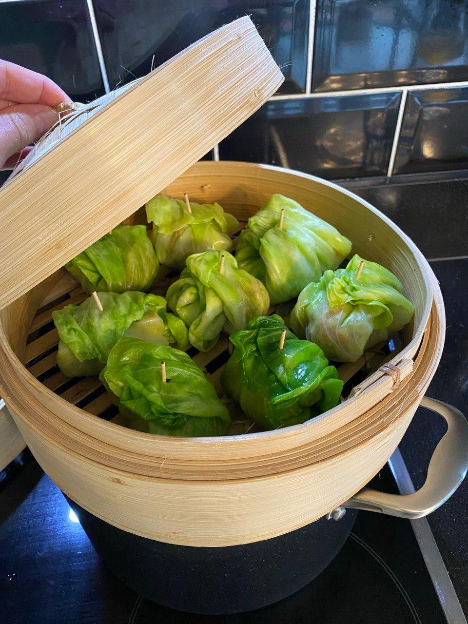 Six steamed cabbage leaf dumplings secured with toothpicks and arranged in a round bamboo steamer basket, viewed from above with the lid lifted by a hand