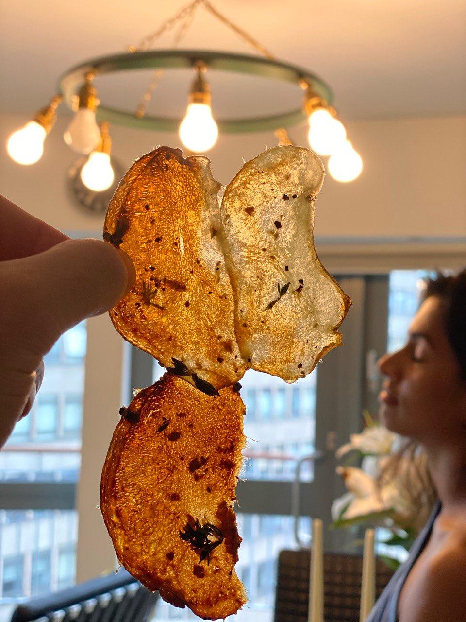 A hand holding up three wafer-thin, golden-brown homemade potato crisps flecked with dark herbs, backlit by a circular pendant light with exposed bulbs in a bright modern flat.