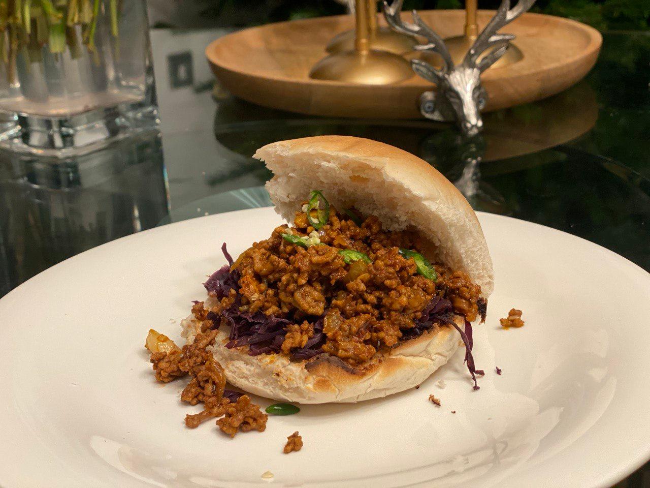 A brioche bun split open and filled with saucy Indian turkey keema, shredded red cabbage and sliced green chilli, served on a white plate on a dark glass table