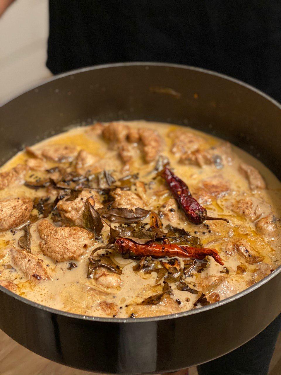 A large dark frying pan held by a person in a black top, filled with pieces of chicken simmering in a golden coconut milk sauce, scattered with fried curry leaves, black mustard seeds and two whole dried red chillies