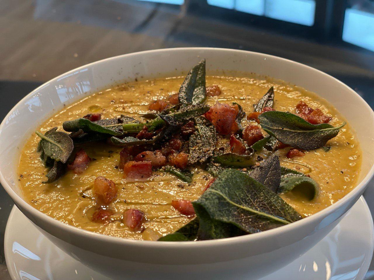 A wide white bowl of velvety golden butternut squash soup, topped with dark-green crispy fried sage leaves and scattered cubes of crispy pancetta, photographed close up on a dark surface