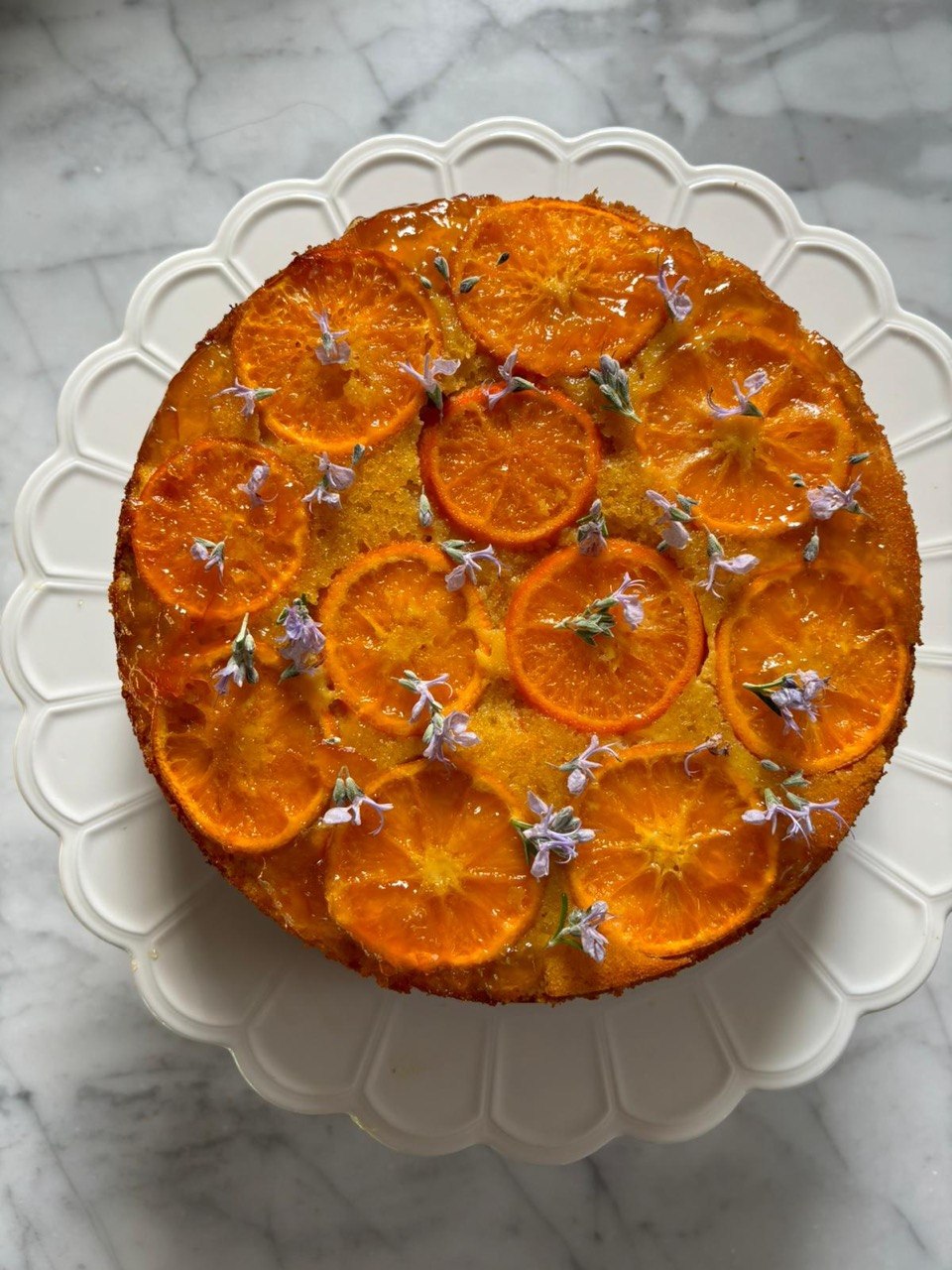 Overhead view of a round upside-down cake on a white scallop-edged cake stand against a marble surface. The top is covered with overlapping caramelised clementine rounds glazed in shiny marmalade, with small purple edible flowers scattered across the surface.