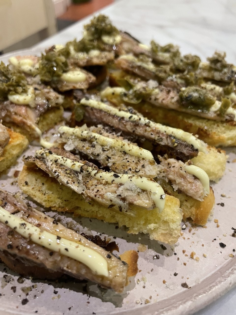 Several slices of toasted sourdough on a white speckled plate, topped with anchovy fillets, a drizzle of pale yellow aioli, cracked black pepper, flaky salt and spoonfuls of vivid green salsa verde