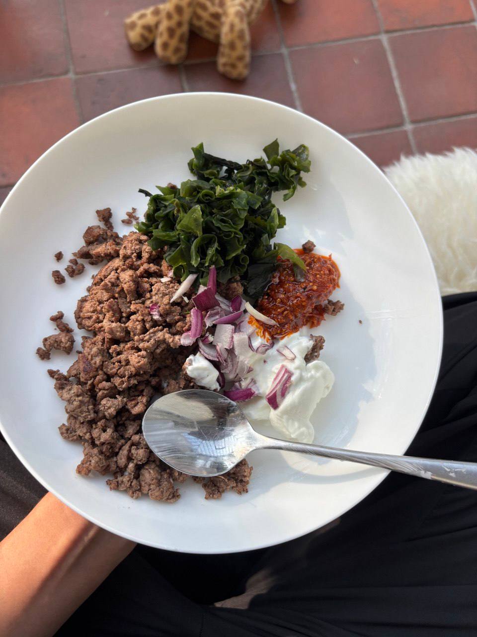 A wide white bowl seen from above, containing browned crumbled beef mince on the left, a pile of dark green rehydrated wakame seaweed at the top, chopped raw red onion in the centre, a dollop of white yogurt and a spoonful of red harissa paste on the right, with a silver spoon resting in the bowl.