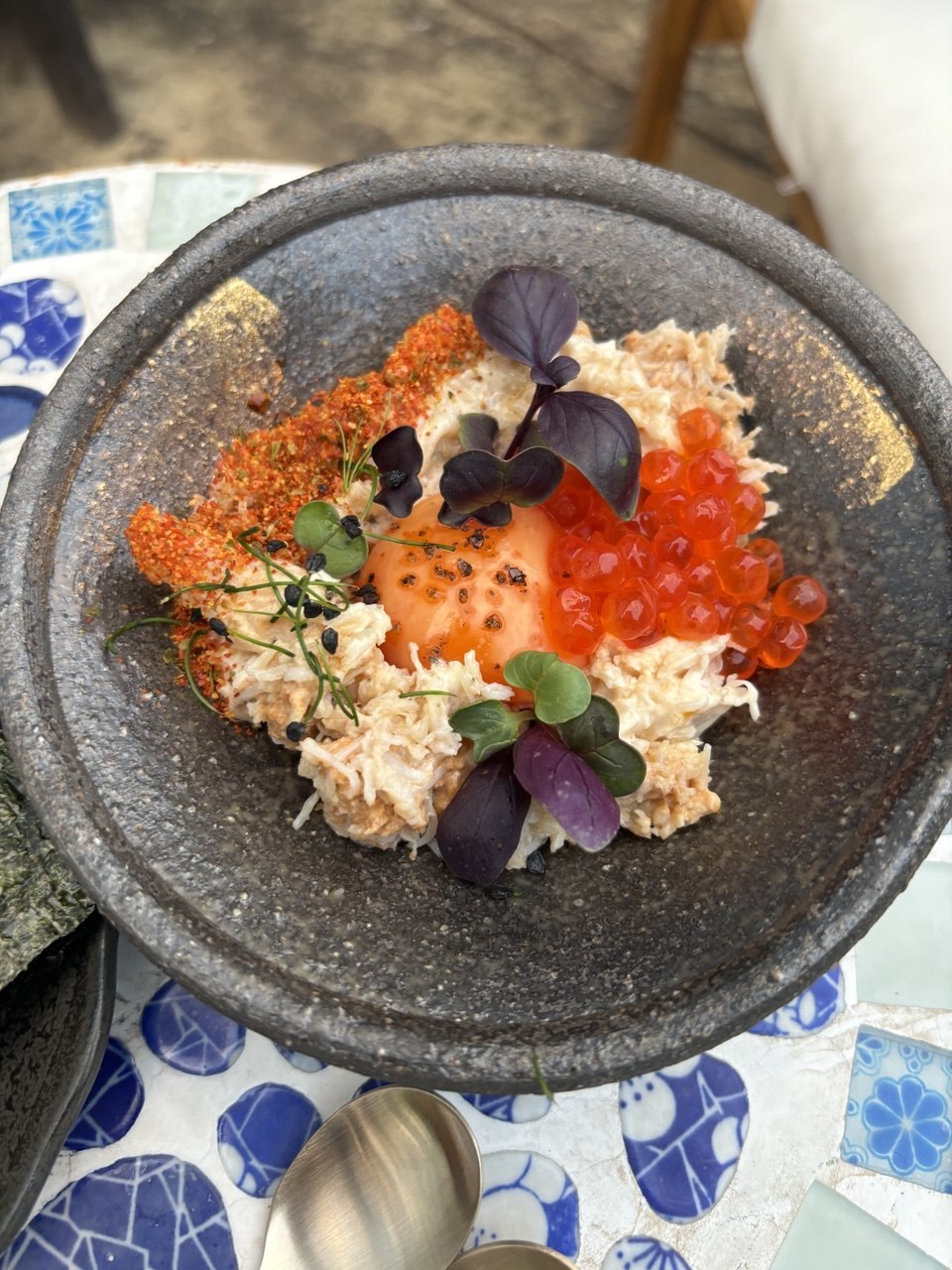 An overhead shot of a dark stone bowl filled with seasoned rice topped with a generous mound of dressed white crab meat, a glossy amber-coloured cured egg yolk in the centre, a cluster of bright red salmon roe, a dusting of red shichimi togarashi spice, and scattered purple basil and green micro herb leaves. The bowl sits on a blue-and-white floral patterned surface with a small metal cup visible at the bottom edge.