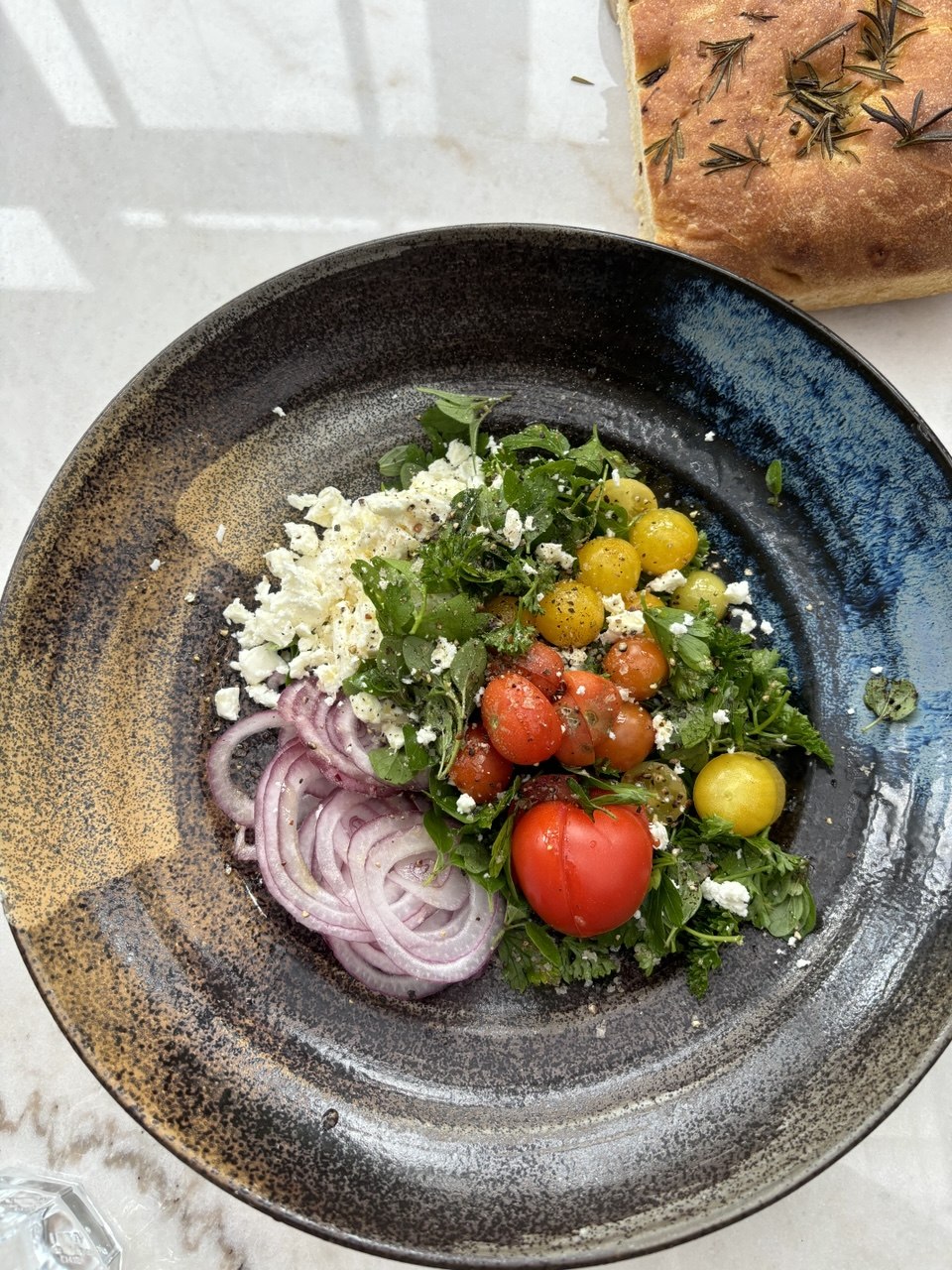 An overhead view of a dark ceramic bowl filled with a salad of red and yellow heirloom tomatoes, crumbled white feta, sliced red onion rings and peppery green herb leaves, served on a marble surface alongside a piece of rosemary focaccia in the top right corner.