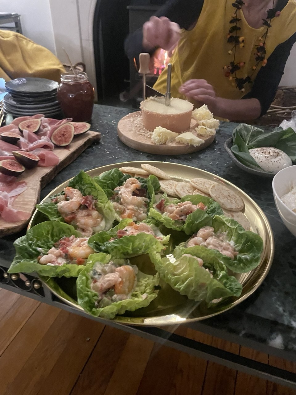 A gold serving platter holding eight little gem lettuce cups each filled with a creamy prawn cocktail mixture. Plain water crackers are tucked to one side. In the background sits a charcuterie board with prosciutto and fresh figs, a whole cheese being flambéed, and a burrata on a dark plate.