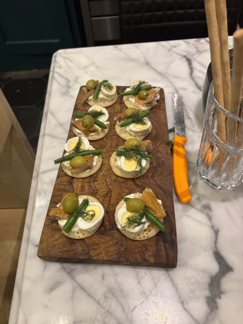 Eight buckwheat blinis arranged on a rectangular wooden board, each topped with light cream cheese, a halved quail egg, a piece of smoked salmon, a green olive, a blanched green bean and a frond of fresh dill, photographed on a marble kitchen counter.