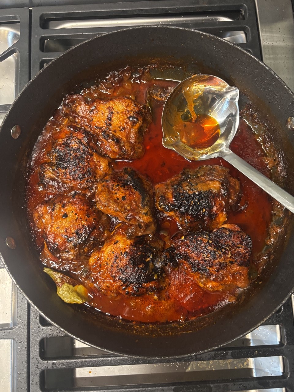 Overhead view of five deeply charred, spiced chicken thighs in a rich, dark red tomato braising sauce inside a large black pan on a gas hob, with a metal ladle resting in the sauce.