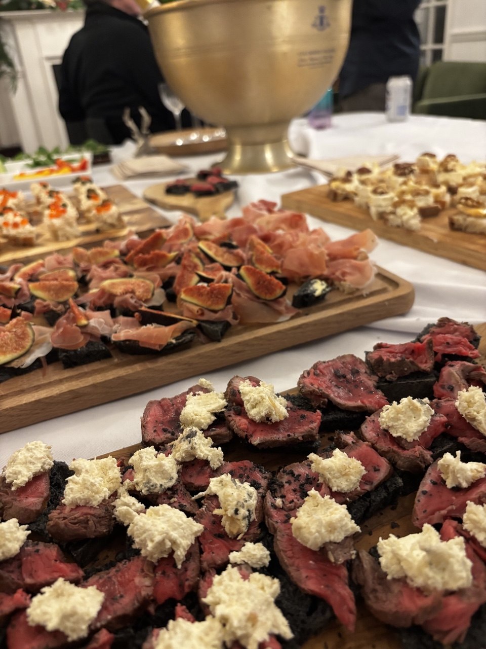 Two wooden serving boards on a white tablecloth: the foreground board is covered in thin slices of pink medium-rare beef on dark crostini, each topped with crumbled white blue cheese; the board behind holds dark toasts topped with folded slices of prosciutto and wedges of fresh fig. A gold champagne bucket and further canapé boards are visible in the background.